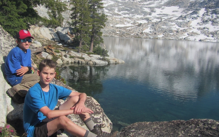 two outward bound students rest on rocks beside a blue alpine lake reflecting a snowy, rocky landscape